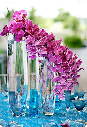 Trailing centerpiece of purple phalaenopsis orchids on top of cylinders with blue glasses