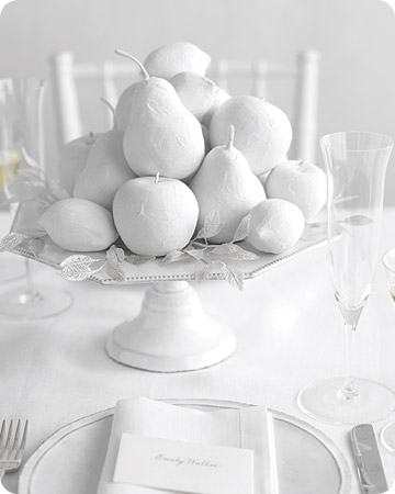 White painted fruit with silver millinery leaves sits atop of a ceramic white cake stand for a monochromatic look;